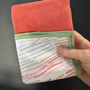May include: Folded coral-colored microfiber cleaning cloth with a patterned bottom section. The bottom section features a white background with colorful diagonal stripes in orange, green, and blue. The edge is trimmed with green fabric.