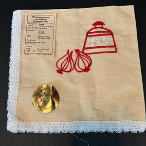 May include: A beige linen tea towel with a red embroidered design of a garlic bulb and a red and white jar with a lid. The text on the label reads "VEB Tapisseriestickerel Am Aschberg" Klingenthal 4, 9653. Erzeugnis Dessin Decke 13-3512-19-NB Scil. Nr.ELN 164 37 620 Material Oweb PAO PEV-Nr. 19 der AO-Nr. Pr. 258/1 Produktion Schlüssel-Nr. 486 12 11 Binnenhandel L. Wahl Größe 40x40 cm eck. EVP M 7,90 3/138