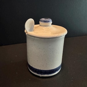 May include: A ceramic jar with a lid and small spoon. The jar is cylindrical with a speckled, off-white finish and a dark blue band at the base. The lid and knob are also off-white with blue accents.