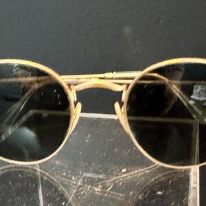 Pu&ograve; includere: Occhiali da sole con montatura dorata e lenti scure. Gli occhiali da sole hanno una classica forma rotonda e una sottile montatura dorata. Il logo Ray-Ban &egrave; visibile sulla lente sinistra. Gli occhiali sono esposti su una superficie trasparente.