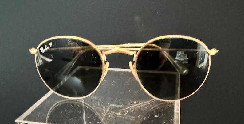 Pu&ograve; includere: Occhiali da sole con montatura dorata e lenti scure. Gli occhiali da sole hanno una classica forma rotonda e una sottile montatura dorata. Il logo Ray-Ban &egrave; visibile sulla lente sinistra. Gli occhiali sono esposti su una superficie trasparente.