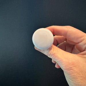 May include: A round, white object is held between fingers against a dark background. The object appears to be smooth and opaque, with a slightly off-white color. The image is a close-up, focusing on the object and the hand.