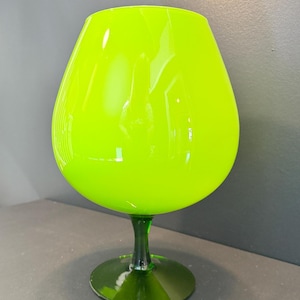 May include: A neon green brandy snifter glass. The glass has a rounded bowl shape and sits on a short stem and a circular base. The glass is made of a shiny, transparent material.