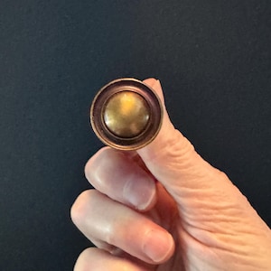 May include: A round, antique-style cabinet knob with a brass-colored center and a bronze-colored outer ring. The knob is held against a dark background, showcasing its metallic finish and circular design.