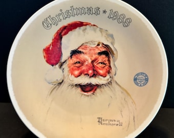May include: A decorative plate featuring a smiling Santa Claus with a red hat and white beard. The plate has the text "Christmas * 1988" and "Norman Rockwell". A small blue emblem is also visible.