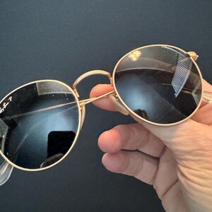 Pu&ograve; includere: Occhiali da sole con montatura dorata e lenti rotonde e scure. Gli occhiali da sole hanno un design classico con una sottile montatura dorata, naselli trasparenti e aste curve. Il logo Ray-Ban &egrave; visibile sulla lente.