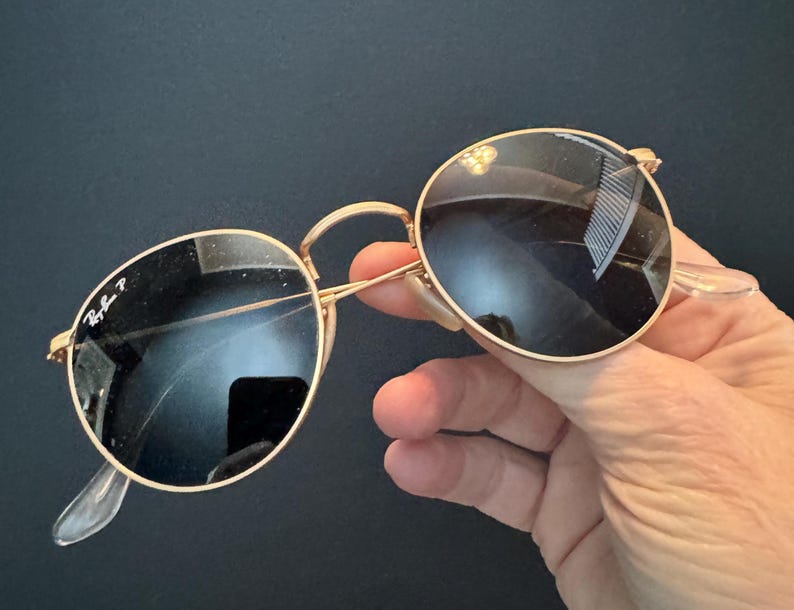 Pu&ograve; includere: Occhiali da sole con montatura dorata e lenti rotonde e scure. Gli occhiali da sole hanno un design classico con una sottile montatura dorata, naselli trasparenti e aste curve. Il logo Ray-Ban &egrave; visibile sulla lente.