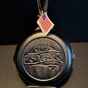 以下が含まれることがあります： 黒い鋳鉄製のフライパン。取っ手と、アメリカ国旗の小さなタグが付いています。「75 YEARS 1938-2013」の文字が底にエンボス加工されており、記念品であることを示しています。