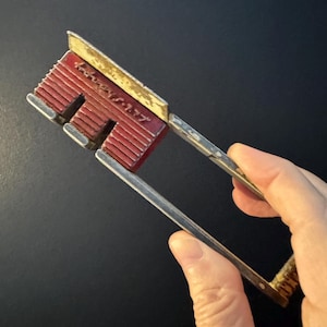 May include: A vintage metal tube wringer tool featuring a red, ribbed, rectangular head with the embossed text "General Slicing" and a long, silver-toned, U-shaped handle. The metal shows signs of wear, including chipped paint and surface oxidation.