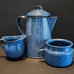 May include: A blue speckled enamelware coffee pot with a matching lid, and two small mugs. The set is a classic camping or rustic kitchenware design. The coffee pot has a handle and spout, and the mugs have handles.