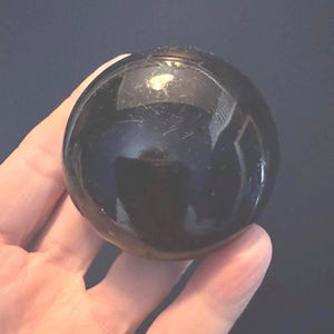 May include: A glossy, black sphere, held in a hand. The surface of the sphere reflects light, creating highlights. The object appears to be made of a solid, polished material. The sphere's smooth surface contrasts with the texture of the hand.