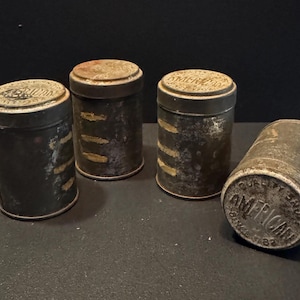 May include: Four cylindrical metal tins with a weathered, dark gray finish. Three tins stand upright, while one lies on its side. Each lid features the embossed text "AMERICAN" and "QUALITY FRUIT" in a circular pattern with a worn, rustic texture.