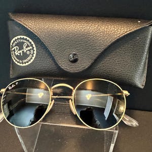 Pu&ograve; includere: Occhiali da sole rotondi con montatura dorata e lenti scure, accompagnati da una custodia in pelle nera. La custodia ha una chiusura a bottone e il logo Ray-Ban. Design classico.