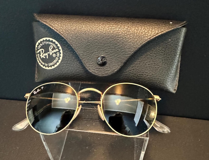 Pu&ograve; includere: Occhiali da sole rotondi con montatura dorata e lenti scure, accompagnati da una custodia in pelle nera. La custodia ha una chiusura a bottone e il logo Ray-Ban. Design classico.
