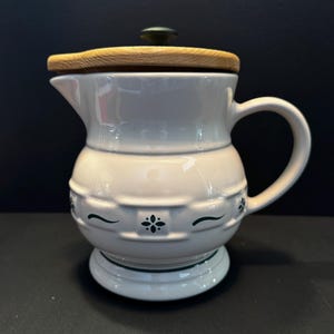 May include: A white ceramic pitcher with a light wood lid and a dark green knob. The pitcher features a rounded body with a textured, embossed pattern around the center, accented by small dark green floral motifs and curved line details.