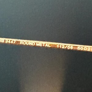 Pu&ograve; includere: Primo piano dell'asta di un paio di occhiali in metallo color oro. Il testo "RB 3447 ROUND METAL 112/58 50[]21 145 3P" &egrave; stampato sull'asta.