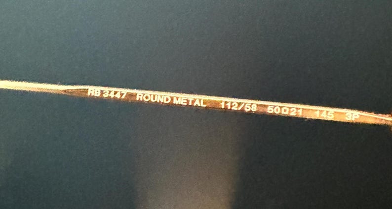 Pu&ograve; includere: Primo piano dell'asta di un paio di occhiali in metallo color oro. Il testo "RB 3447 ROUND METAL 112/58 50[]21 145 3P" &egrave; stampato sull'asta.
