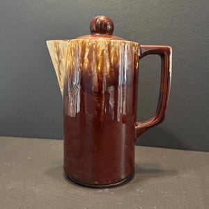 May include: A vintage brown ceramic coffee pot with a lid and handle. The pot has a glossy finish with a dripped glaze effect in shades of brown and tan. The handle is a dark brown color.