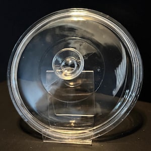 May include: A clear glass lid with a circular shape and a small, round handle in the center. The lid has a slightly raised rim and is designed to fit on a pot or pan. The lid is displayed on a clear plastic stand.