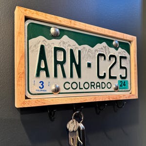May include: A Colorado license plate key holder with a natural wood frame. The license plate is green and white with the text "ARN-C25 COLORADO". Includes four black hooks for keys and a key with a dog-shaped tag.