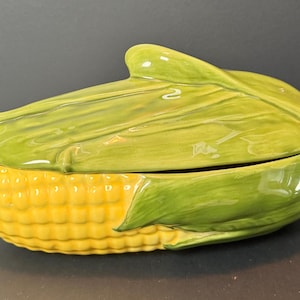 May include: A ceramic corn-shaped serving dish with a lid. The dish is yellow with a textured surface, mimicking corn kernels. The lid is green, resembling corn husks. The dish is designed to hold food and is likely used for serving.