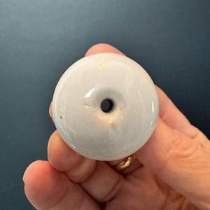 May include: A light gray ceramic donut-shaped bead with a central hole. The bead has a glossy finish and a small gold accent. The object is held in a hand against a dark background.