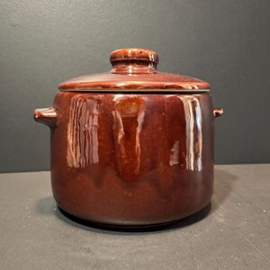 May include: A brown ceramic lidded pot with two side handles. The pot has a glossy finish and a rounded shape. The lid has a small knob on top. The pot is sitting on a dark surface.