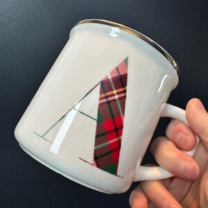 May include: White ceramic mug with a gold rim and a handle. The letter "A" is prominently displayed on the front, featuring a red, green, and brown plaid pattern. The mug is held against a dark background.
