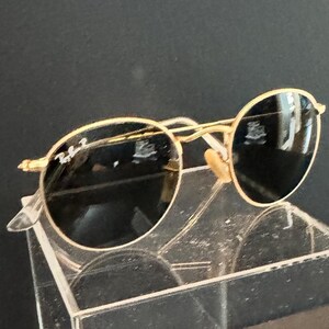 Pu&ograve; includere: Occhiali da sole con montatura dorata e lenti rotonde. Gli occhiali da sole hanno un design classico con una sottile montatura dorata e lenti scure. Il logo Ray-Ban &egrave; visibile sulla lente sinistra. Gli occhiali sono esposti su un supporto in acrilico trasparente.