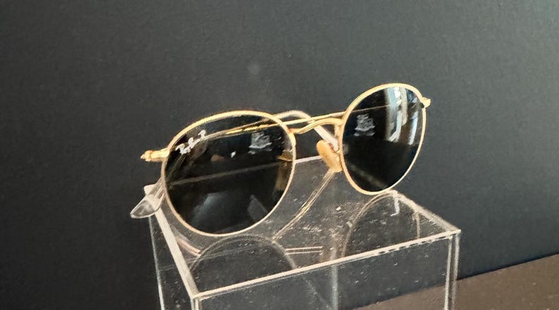 Pu&ograve; includere: Occhiali da sole con montatura dorata e lenti rotonde. Gli occhiali da sole hanno un design classico con una sottile montatura dorata e lenti scure. Il logo Ray-Ban &egrave; visibile sulla lente sinistra. Gli occhiali sono esposti su un supporto in acrilico trasparente.