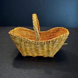 May include: A small, oval-shaped woven basket with a light golden-brown finish. The basket features a single, thick, braided handle that arches over the center, and the sides are constructed with a horizontal weave pattern and a scalloped rim.