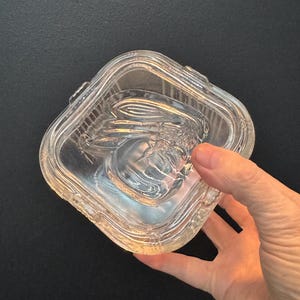 May include: A clear, square glass dish with a textured design and a lid. The dish has a decorative pattern on the inside and is held by a hand against a dark background. The dish is likely for serving or storage.