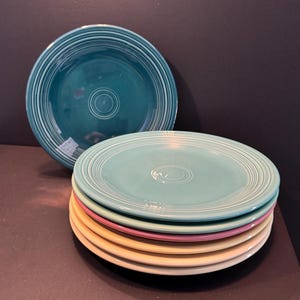 May include: A stack of colorful ceramic plates in shades of teal, light blue, pink, and yellow. The plates have concentric ring designs and a central indentation. One teal plate is positioned behind the stack.