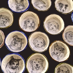 Puede incluir: Una colección de pequeños cuencos de cerámica redondos, cada uno con una ilustración única en blanco y negro estilo tinta de una cara de perro o gato en el centro. Los cuencos tienen bordes de colores variados, incluyendo azul, verde y marrón.