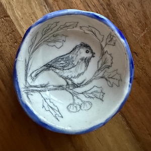 Puede incluir: Pequeño plato redondo de cerámica con un diseño de pájaro y rama dibujado a mano. El pájaro está esbozado en negro sobre un interior blanco, con un borde azul. El plato mide probablemente 10 cm de diámetro.