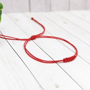 May include: A red string bracelet with a knotted design. The bracelet is tied in a simple knot and is made of a thin, red cord.