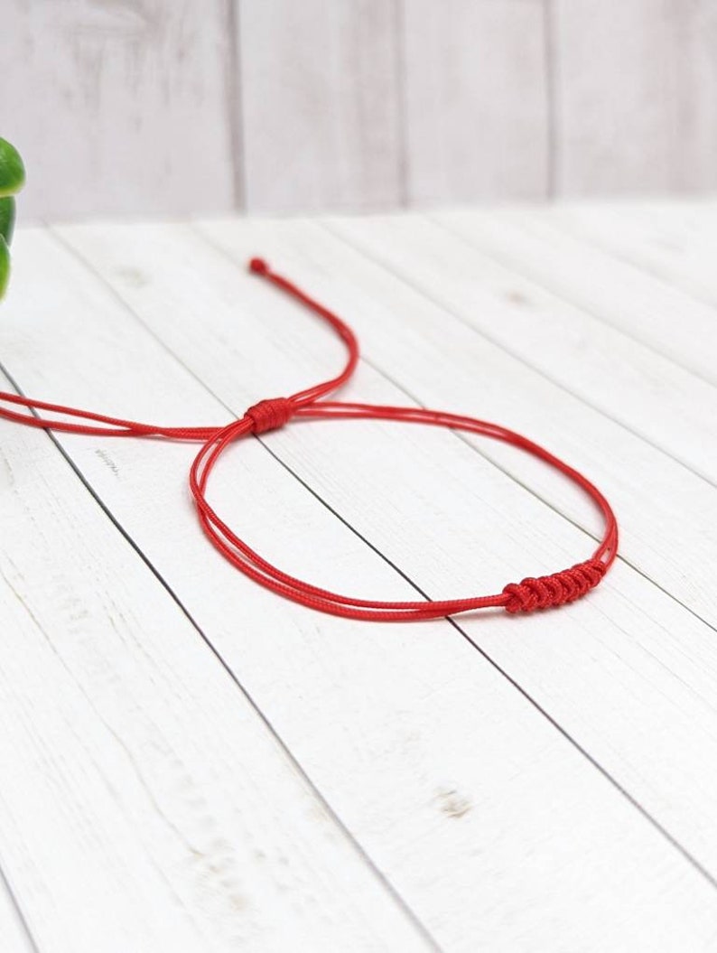 May include: A red string bracelet with a knotted design. The bracelet is tied in a simple knot and is made of a thin, red cord.