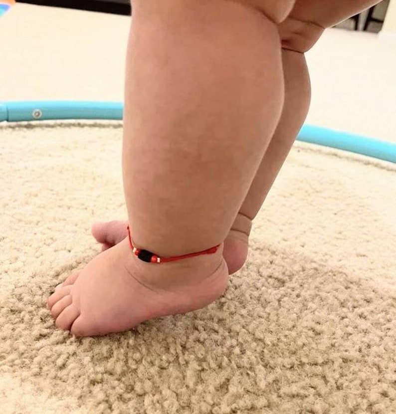 May include: A close-up of a baby's feet with a red string bracelet with a black bead on one ankle.