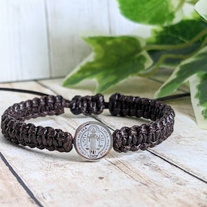 May include: A brown braided bracelet with a silver Saint Christopher medal. The medal features a depiction of Saint Christopher carrying a child on his shoulders. The medal is inscribed with the words "Sanctus Christopherus" and "Protector Viatorum".