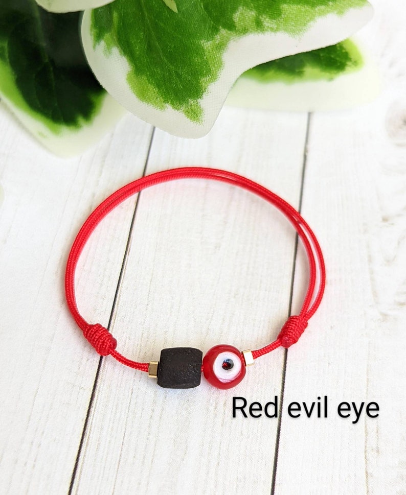 May include: Red string bracelet with a black bead and a red evil eye charm. "Red evil eye" is written below the bracelet.