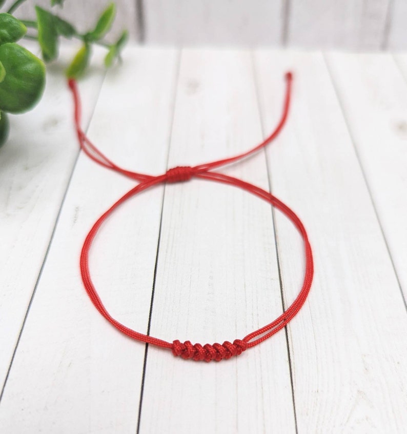 May include: A red string bracelet with a knotted design. The bracelet is tied in a simple knot and is made of a thin, red string.
