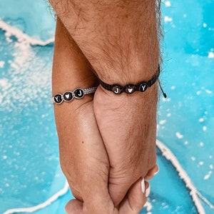 May include: Two people holding hands, each wearing a beaded bracelet with black beads and white letters spelling "J" and "Y" with a heart symbol between the letters.