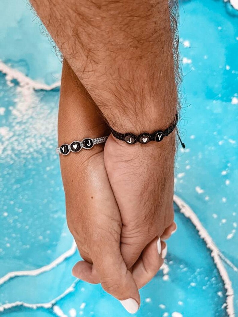 May include: Two people holding hands, each wearing a beaded bracelet with black beads and white letters spelling "J" and "Y" with a heart symbol between the letters.