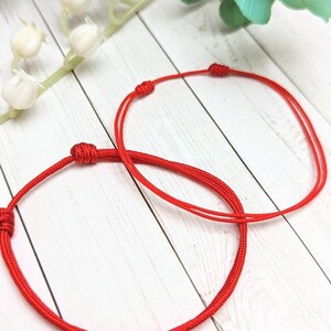 May include: Two red string bracelets tied in knots. The bracelets are on a white wooden surface.