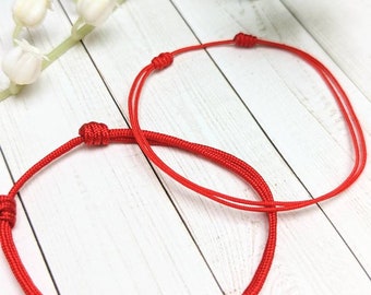 May include: Two red string bracelets tied in knots. The bracelets are on a white wooden surface.