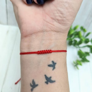 May include: A red string bracelet with a knot in the center. The bracelet is worn on a person's wrist.