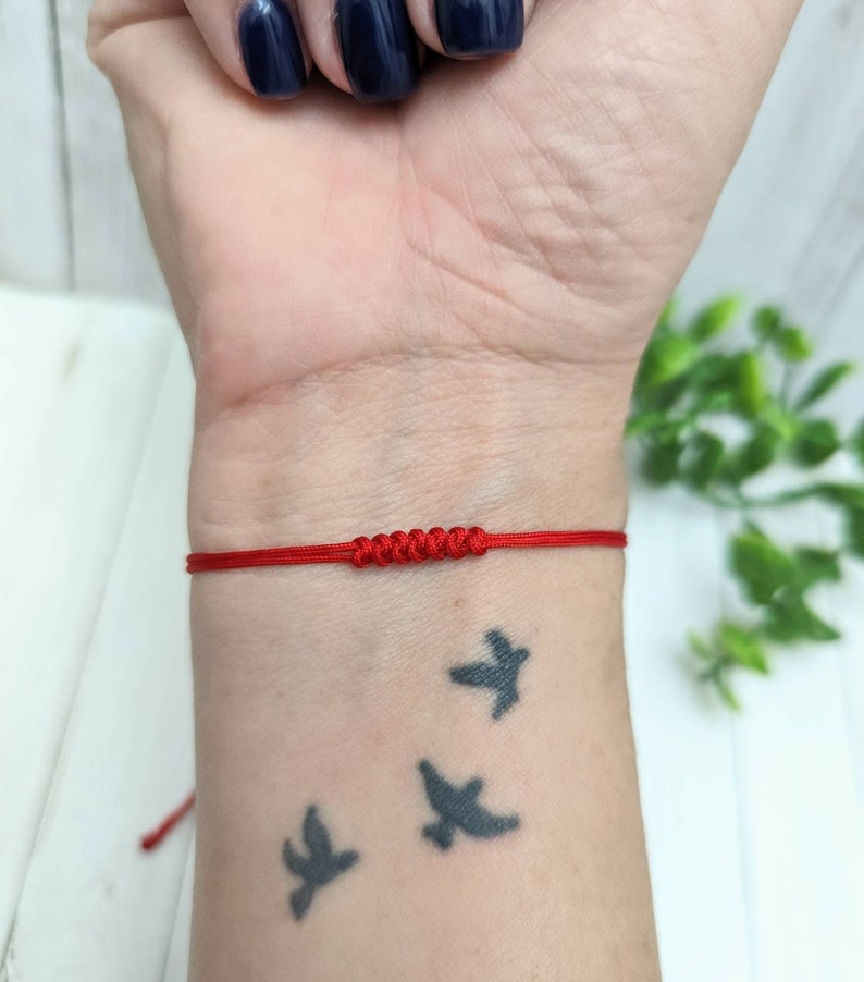 May include: A red string bracelet with a knot in the center. The bracelet is worn on a person's wrist.