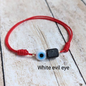 May include: Red string bracelet with a white evil eye charm and a black bead. The bracelet is tied with a knot and the text "White evil eye" is written below the bracelet.