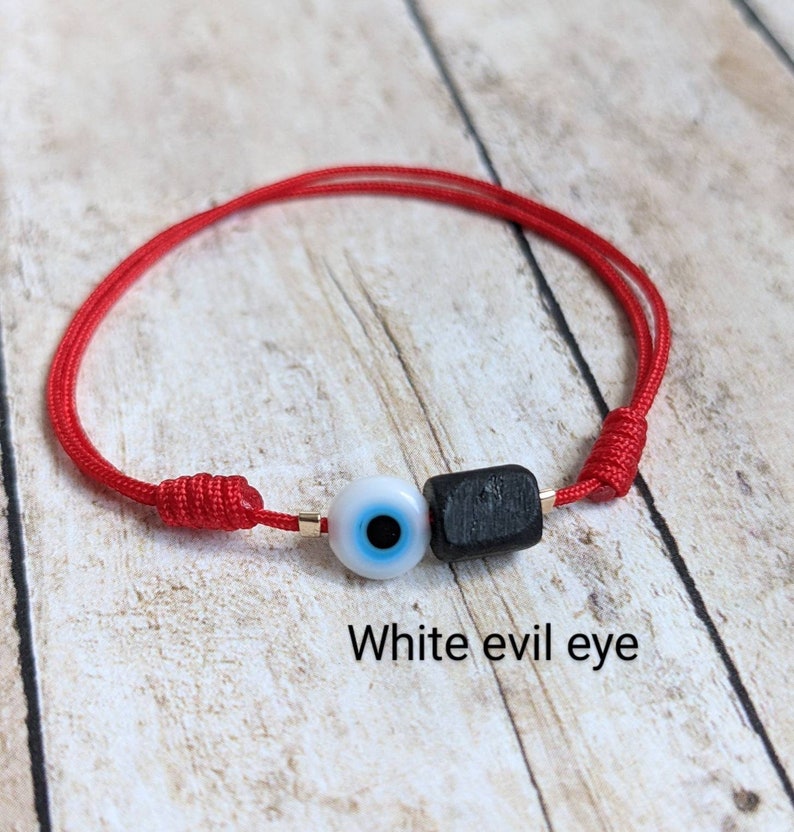 May include: Red string bracelet with a white evil eye charm and a black bead. The bracelet is tied with a knot and the text "White evil eye" is written below the bracelet.