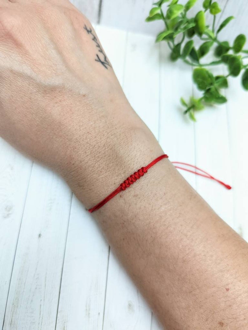 May include: A red string bracelet with a knotted design. The bracelet is tied around a person's wrist.